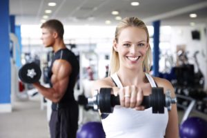 Beautiful young woman smiles at the camera while lifting weights at the gym. Horizontal shot.